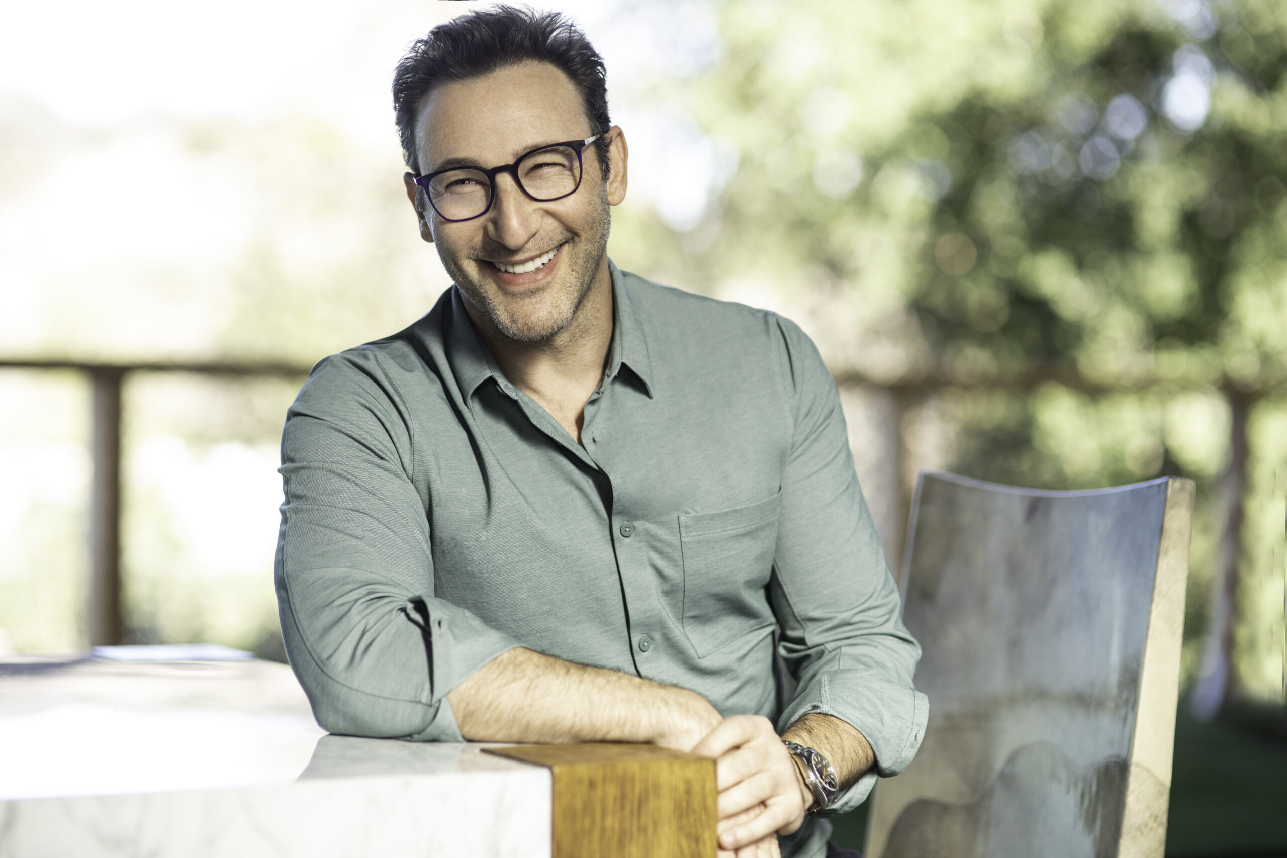 Headshot of Simon Sinek wearing a gray button down shirt, glasses, and leaning on a counter.
