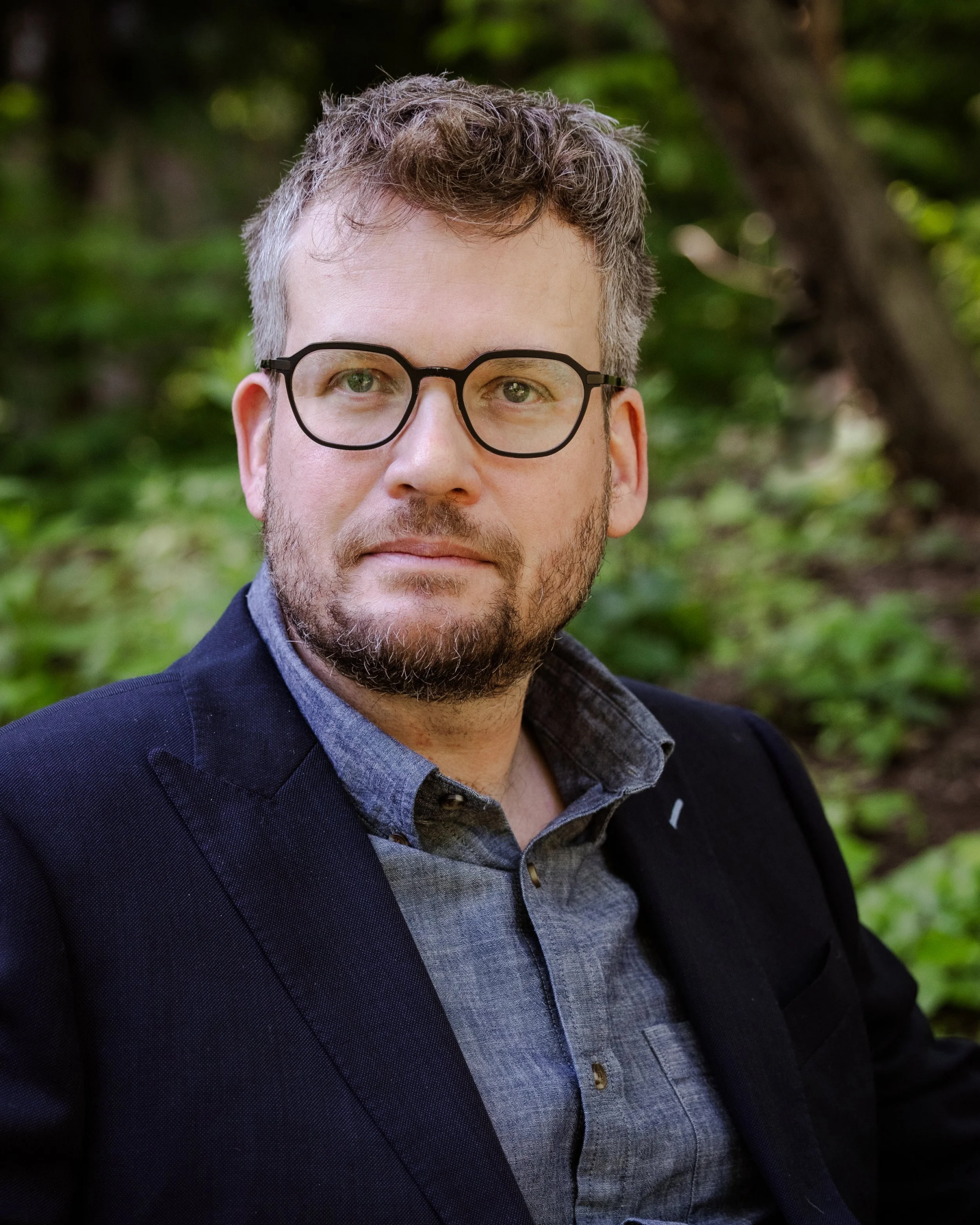 Headshot of John Green, wearing glasses, a button down shirt, and suit jacket.
