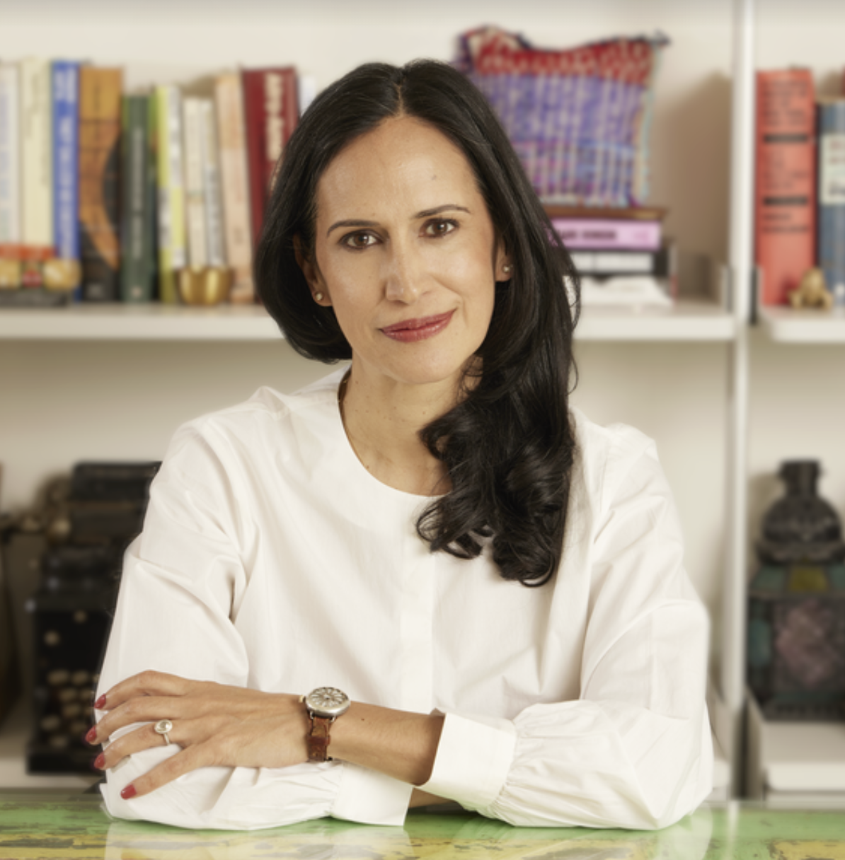 Headshot of Priya Parker sitting at a table, wearing a long sleeved white shirt with her arms crossed. There is a bookshelf behind her with books and art pieces.